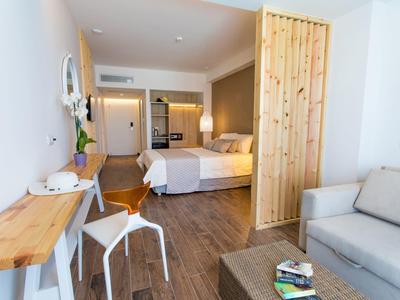 Modernes, helles Zimmer mit hellgrauem Sofa, Holztisch, Bett und Arbeitsbereich mit Hocker.