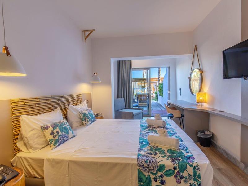 Jasny pokój hotelowy z podwójnym łóżkiem, kolorowymi poduszkami, telewizorem na ścianie i balkonem z miejscem do siedzenia.