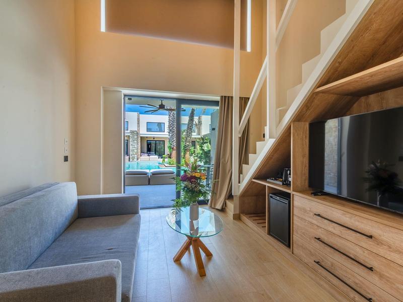 Salon z szarą sofą, szklanym stolikiem i telewizorem pod schodami w nowoczesnym pokoju hotelowym.