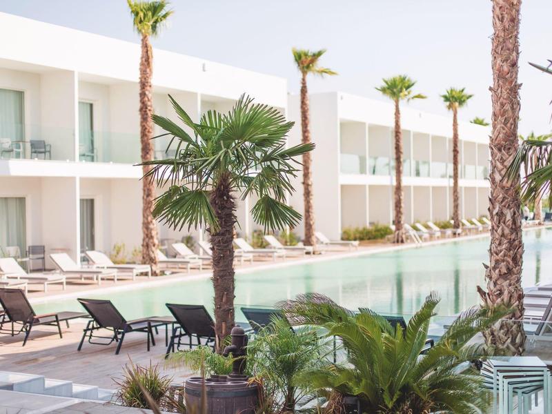 Nowoczesny basen hotelowy z leżakami i palmami w białym kompleksie budynków.