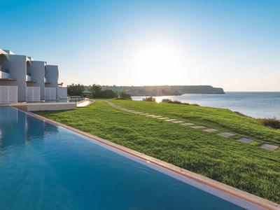 Infinity zwembad met uitzicht op groen gazon en oceaan bij zonsondergang naast moderne witte hotelkamers.