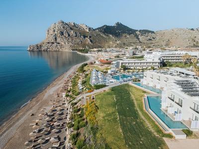 Pohled na hotel s bazénem, pláží a skalnatým kopcem pod jasnou modrou oblohou.
