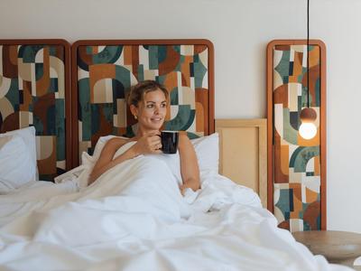 Žena sedí v posteli moderního hotelového pokoje s barevnými geometrickými panely čela postele.