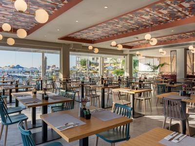 Světla restaurace s dřevěnými stoly, židlemi a velkými okny s výhledem na terasu a moře.