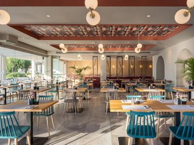 Světla hotelová restaurace s moderními dřevěnými stoly, modrými židlemi, velkými okny a stropním osvětlením.