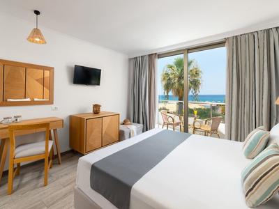 Moderne hotelkamer met tweepersoonsbed, bureau, tv en balkon met uitzicht op palmbomen en oceaan.