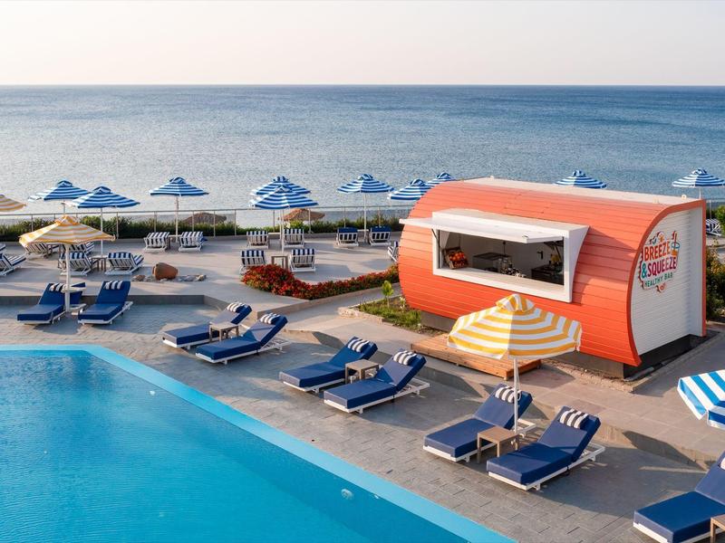 Strand met blauw-witte parasols en ligstoelen bij een zwembad en een kleine oranje kiosk.