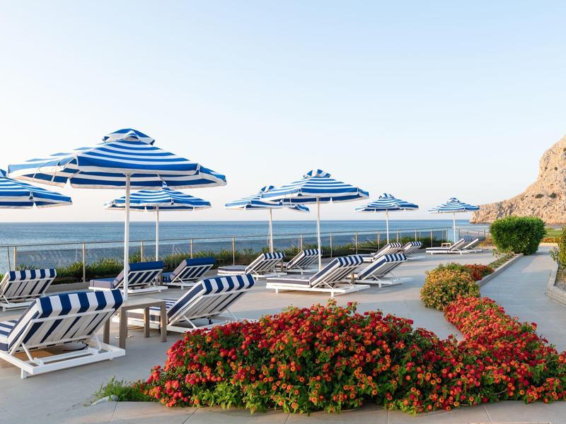 Ligbedden met blauwe parasols naast bloeiende planten met uitzicht op zee.