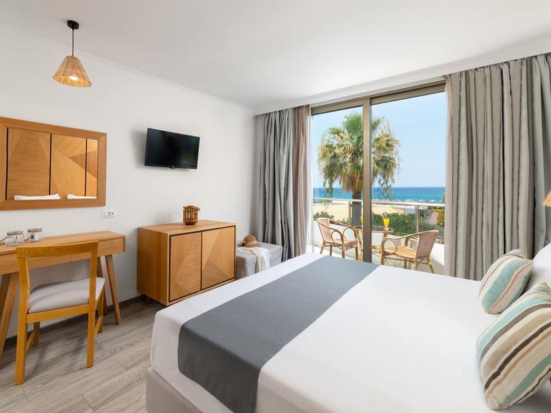 Moderne hotelkamer met tweepersoonsbed, bureau, tv en balkon met uitzicht op palmbomen en oceaan.