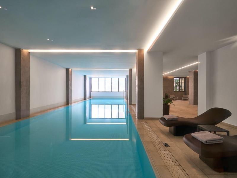 Piscina interna moderna con linee pulite, lettini in legno e grande finestra per luce naturale.