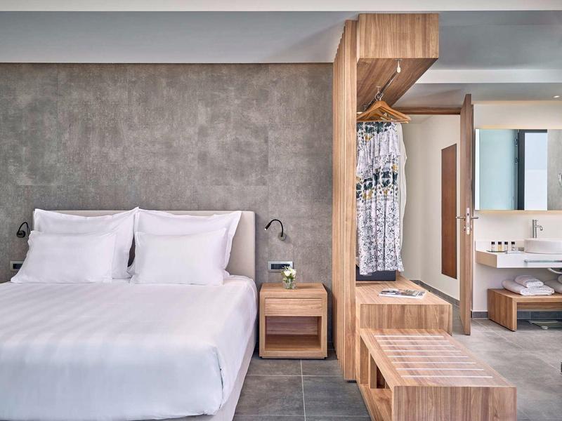 Camera d'hotel moderna e accogliente con letto matrimoniale, comodino e guardaroba aperto in tonalità neutre.