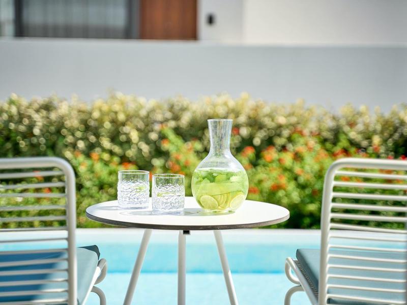 Piccolo tavolo rotondo da esterno con caraffa e bicchieri, circondato da sedie e piante sullo sfondo.