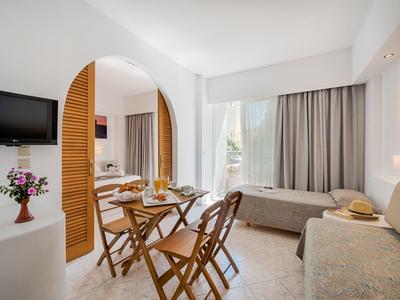 Heller Wohnraum mit Holztisch, Klappstühlen, zwei Sofas und großem Fenster mit Vorhängen.