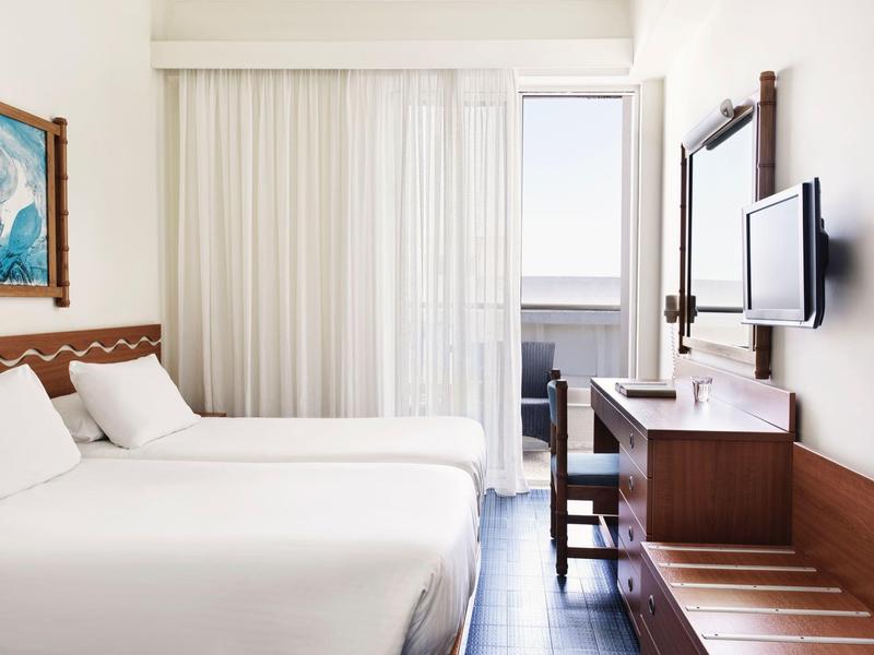 Helles Hotelzimmer mit zwei Betten, weißer Gardine, Schreibtisch, Fernseher und Balkonzugang.