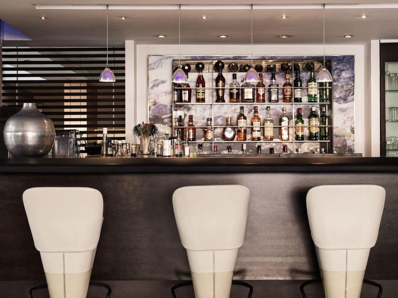 Drei weiße Barhocker vor einer schwarzen Bar mit beleckter Regalwand und Spirituosen.