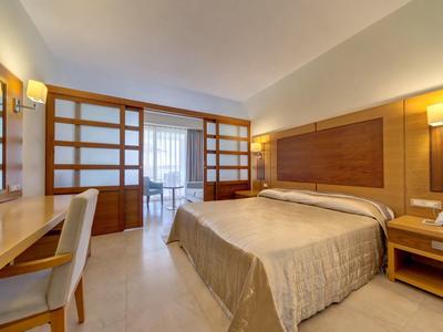 Habitación de hotel luminosa con cama doble, escritorio, silla y detalles de madera, acceso a balcón.