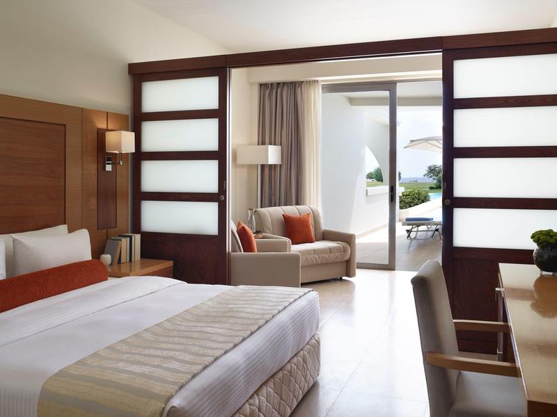 Habitación de hotel moderna con cama grande, sillón, mesa y vista a la terraza con jardín.