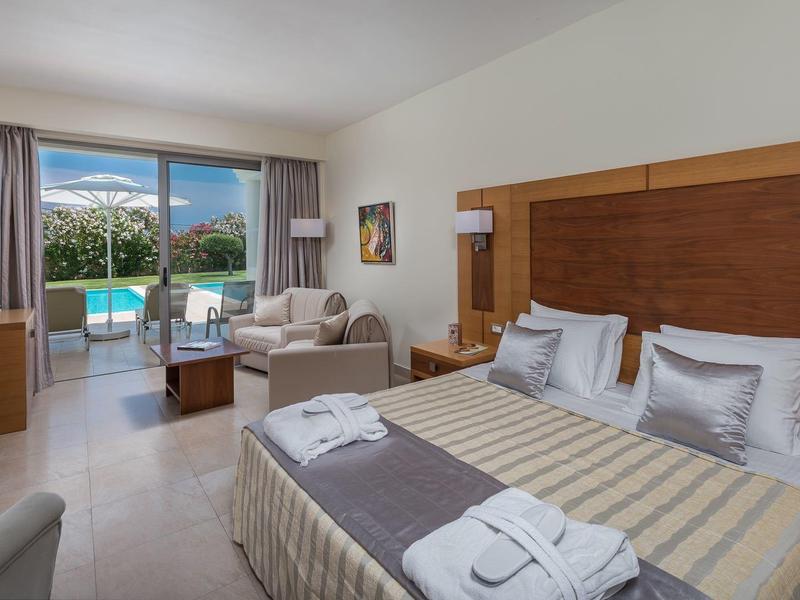 Habitación de hotel luminosa con cama doble, balcón y vista a una piscina y montañas al fondo.