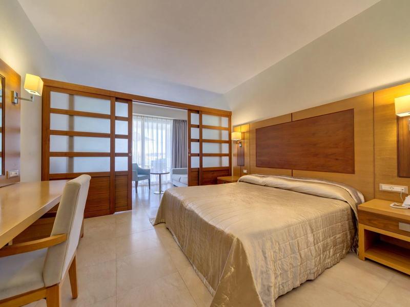 Habitación de hotel luminosa con cama doble, escritorio, silla y detalles de madera, acceso a balcón.