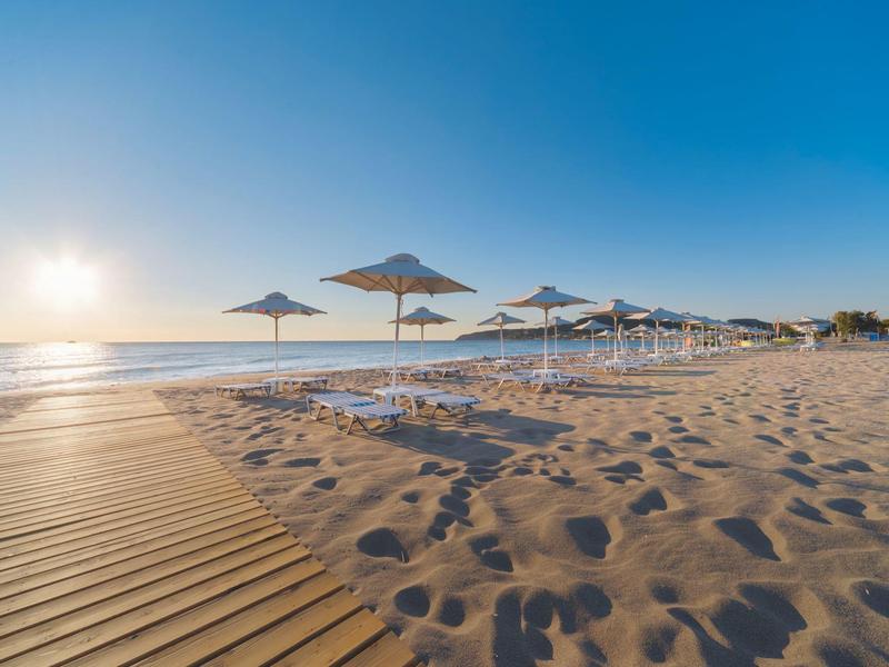 Playa con sombrillas y pasarela de madera al atardecer junto al mar tranquilo.