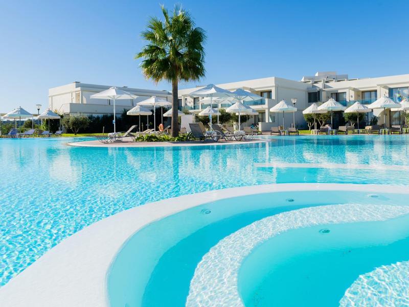 Piscina clara con escalones frente a un hotel moderno y palmeras bajo un cielo despejado.