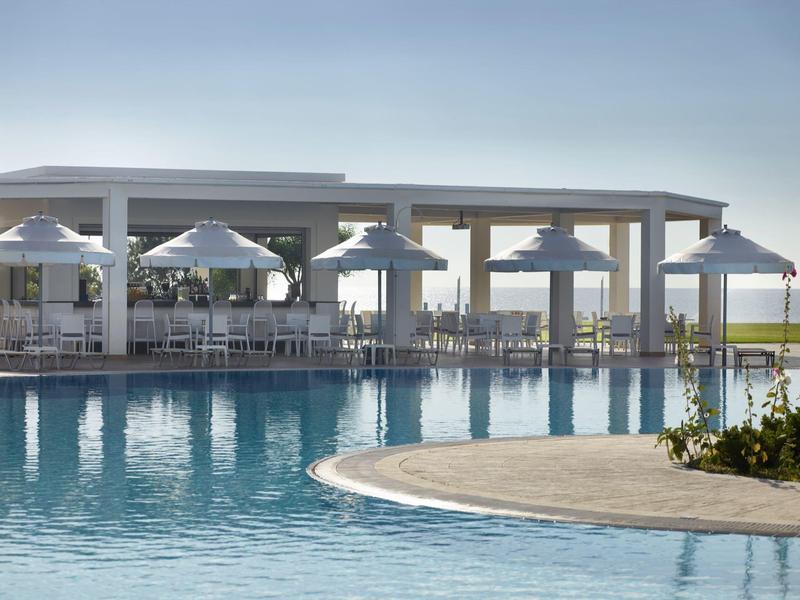 Piscina al aire libre moderna con sombrillas y áreas de descanso, rodeada de un pabellón abierto y cielo azul.