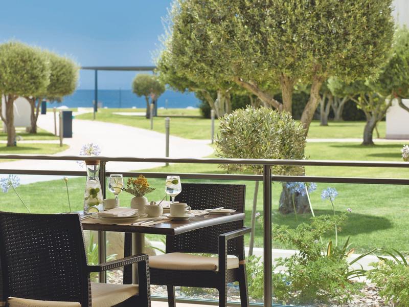 Terraza con mesas y sillas en jardín verde con árboles y vista al mar.