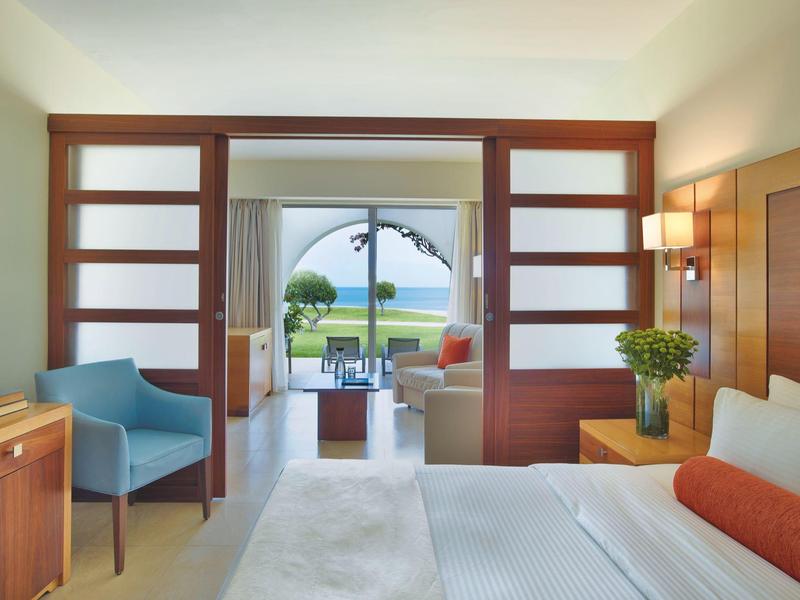 Habitación de hotel moderna con vista al jardín y al mar a través de grandes puertas de vidrio.