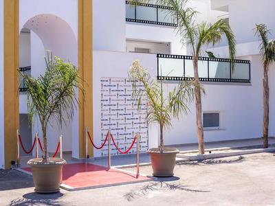 Wejście do hotelu z czerwonym dywanem, barierkami i roślinami doniczkowymi przed białym budynkiem.