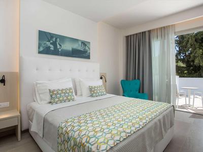 Jasny pokój hotelowy z podwójnym łóżkiem, turkusowym fotelem i balkonem z widokiem na zieleń.