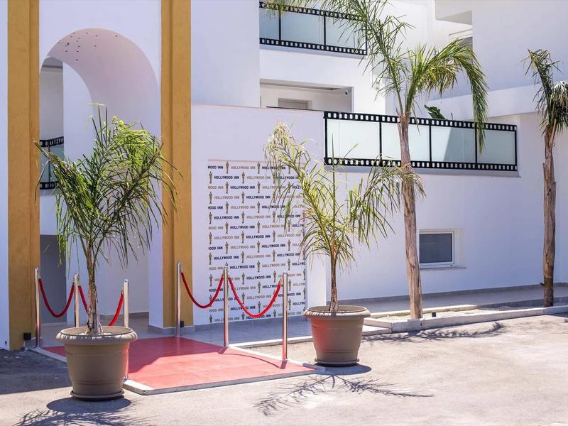 Wejście z czerwonym dywanem, słupkami i palmami przed białym budynkiem hotelu.