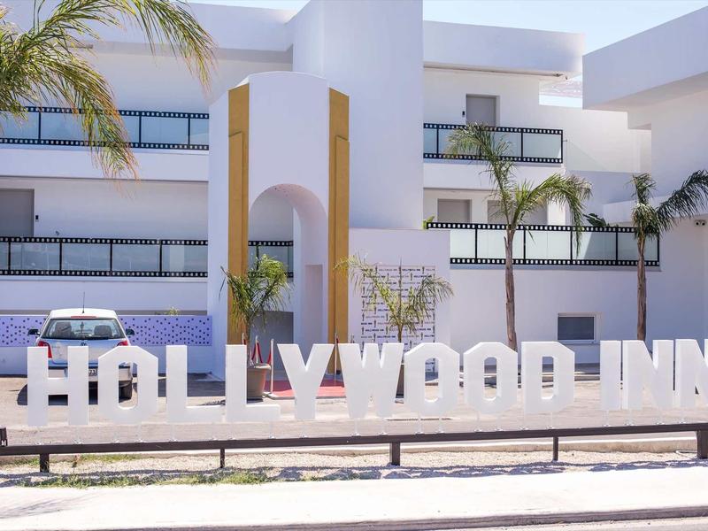 Duży biały napis HOLLYWOOD INN przed nowoczesnym budynkiem z palmami.