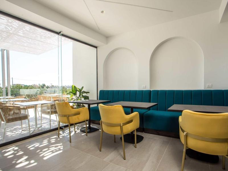 Jasna sala jadalna w hotelu z żółtymi krzesłami, niebieskimi ławkami i tarasem z widokiem na zieleń.