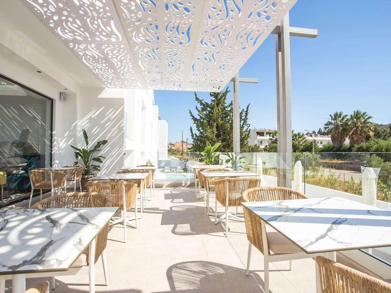 Słoneczny taras z białymi stołami i krzesłami przed hotelem, otoczony roślinami.