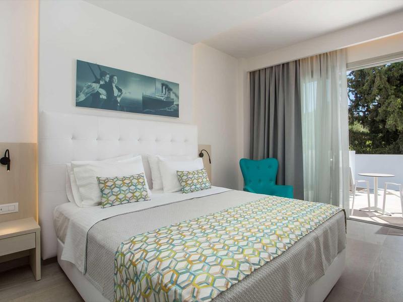 Jasny pokój hotelowy z podwójnym łóżkiem, turkusowym fotelem i balkonem z widokiem na zieleń.