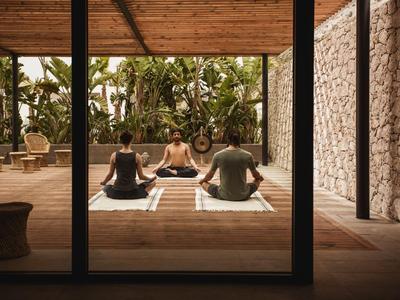 Tre persone meditano su tappetini da yoga in una stanza aperta con vista sulle piante.