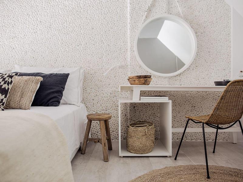 Lichte, minimalistische slaapkamer met witte muren, ronde spiegel en rieten stoel bij een bureau.