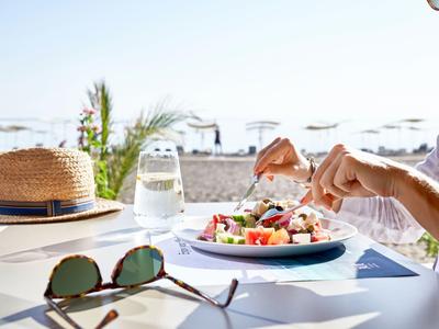Ein gedeckter Tisch mit Salat, Sonnenbrille, Glas Wasser und Strohhut am Strand bei Sonnenschein.