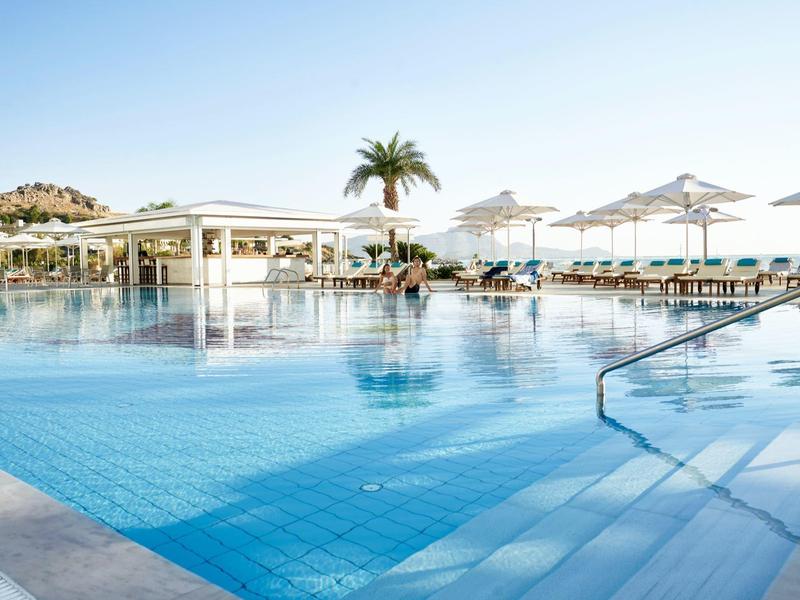 Grande piscine avec eau claire, parasols et un palmier sous un ciel bleu.