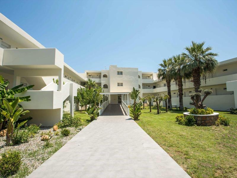 Nowoczesny biały kompleks hotelowy z zadbanym ogrodem i palmami pod niebieskim niebem.