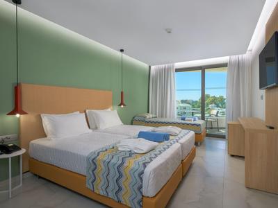 Habitación de hotel moderna con cama doble, cama individual, pared verde y vista al mar.