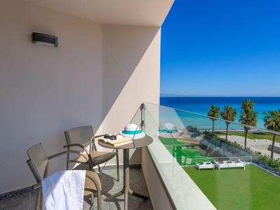 Balcón con mesa y sillas, vista al mar y palmeras bajo un cielo azul.