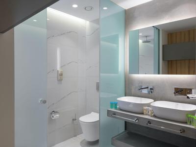 Baño moderno de hotel con ducha de vidrio, inodoro y lavabos dobles en diseño luminoso.