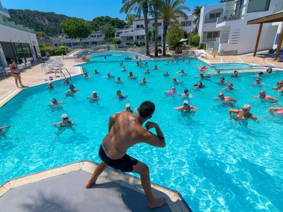 Hombre lidera aeróbicos acuáticos en gran piscina con muchos participantes en un día soleado.