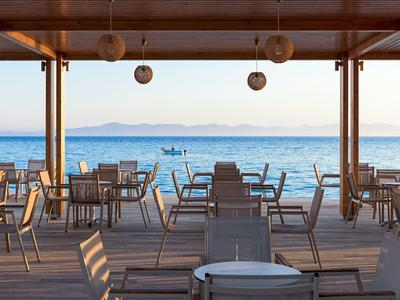 Leere Außenterrasse mit Stühlen und Tischen unter Holzpergola, Blick auf ruhiges Meer und Berge.
