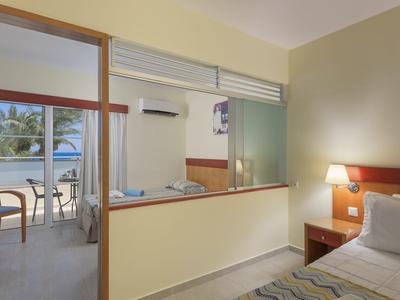 Helles Hotelzimmer mit Einzelbett, Holzmöbeln und Balkon mit Meerblick und Palm Bäumen.