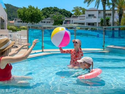 Drei Personen spielen mit einem bunten Strandball im Außenpool an einem sonnigen Tag.