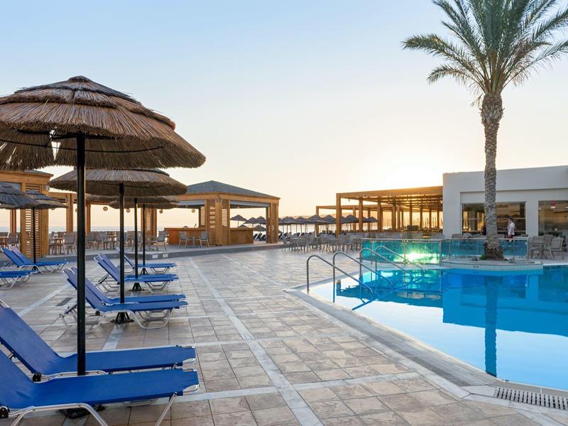 Schwimmbad mit blauen Liegen, Strohdächern, Palmen und moderner Anlage bei klarem Himmel.