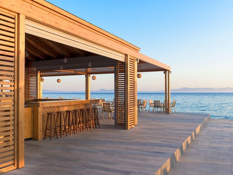 Offene Holzterrasse mit Bar am Meer, blauer Himmel, ruhiges Wasser im Hintergrund.