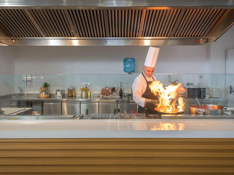 Koch mit weißer Uniform und Hut bereitet flambiertes Gericht hinter Schutzglas in moderner Küche zu.